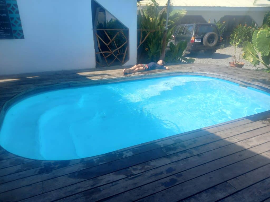 Young boy in swimgear lies on his back on the wooden decking next to a small oval shaped swimming pool