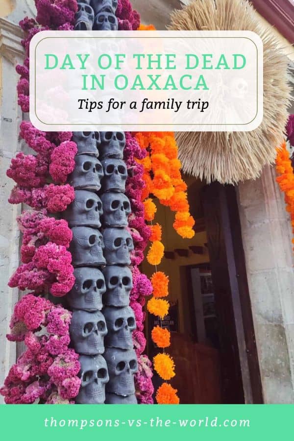 Colorful and festive image of a doorway in Oaxaca adorned with a garland of pink flowers and black skulls, complemented by orange marigold decorations and a straw ornament, celebrating Day of the Dead. Text overlay reads "DAY OF THE DEAD IN OAXACA - Tips for a family trip" in green and white. Perfect for families planning a Day of the Dead experience in Oaxaca! Save this pin to your Pinterest board for later reference.