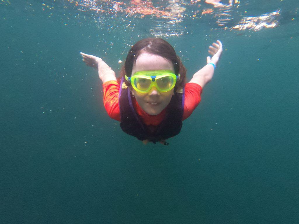 Young boy swims under water in a cenote, wearing goggles and with his arms stretched behind him