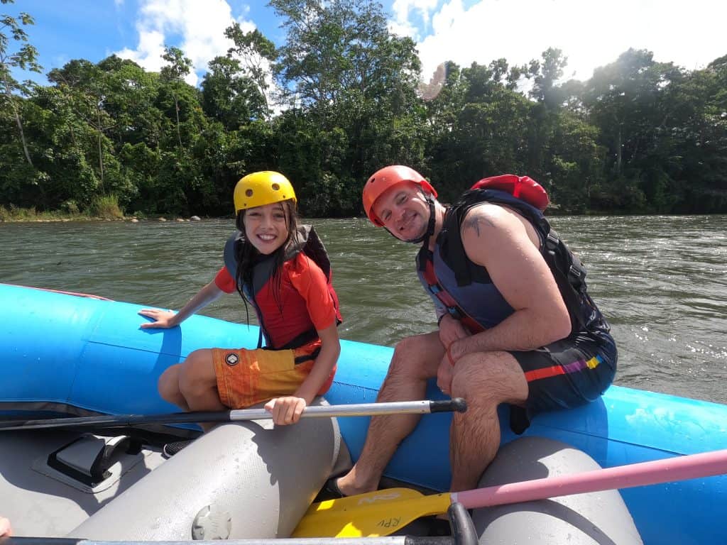 Young boy and man in swim gear, life jackets and helmets sit on edge of blue inflatable raft with flowing river in the background
