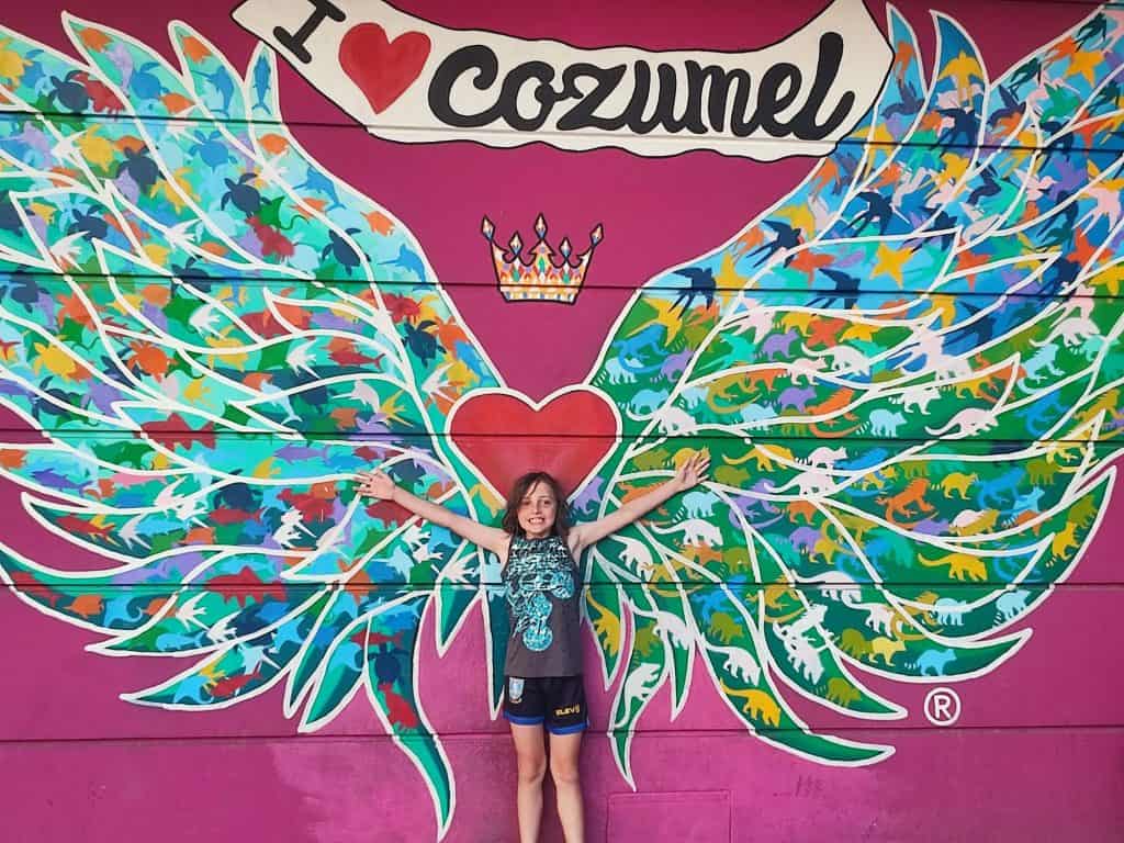 Boy stands in front of painted 'I Love Cozumel' sign on a wall with his arms outstretched