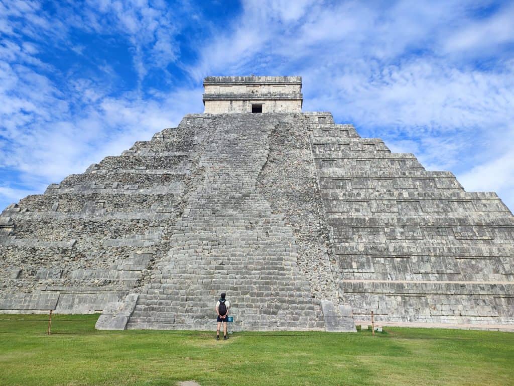 Man stands at the base of El Castillo, the giant Mayan pyramid at the centre of Chichen Itza in Mexico. He is wearing a backpack and looking upwards to the top of the structure