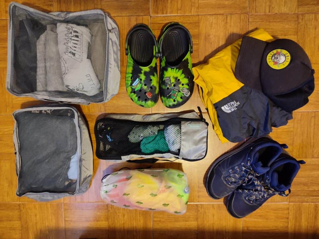 Kids clothes lie on floor in packing cubes of various sizes, alongside a pair of green tie dye crocs, some hiking trainers and a Guns n Roses cap