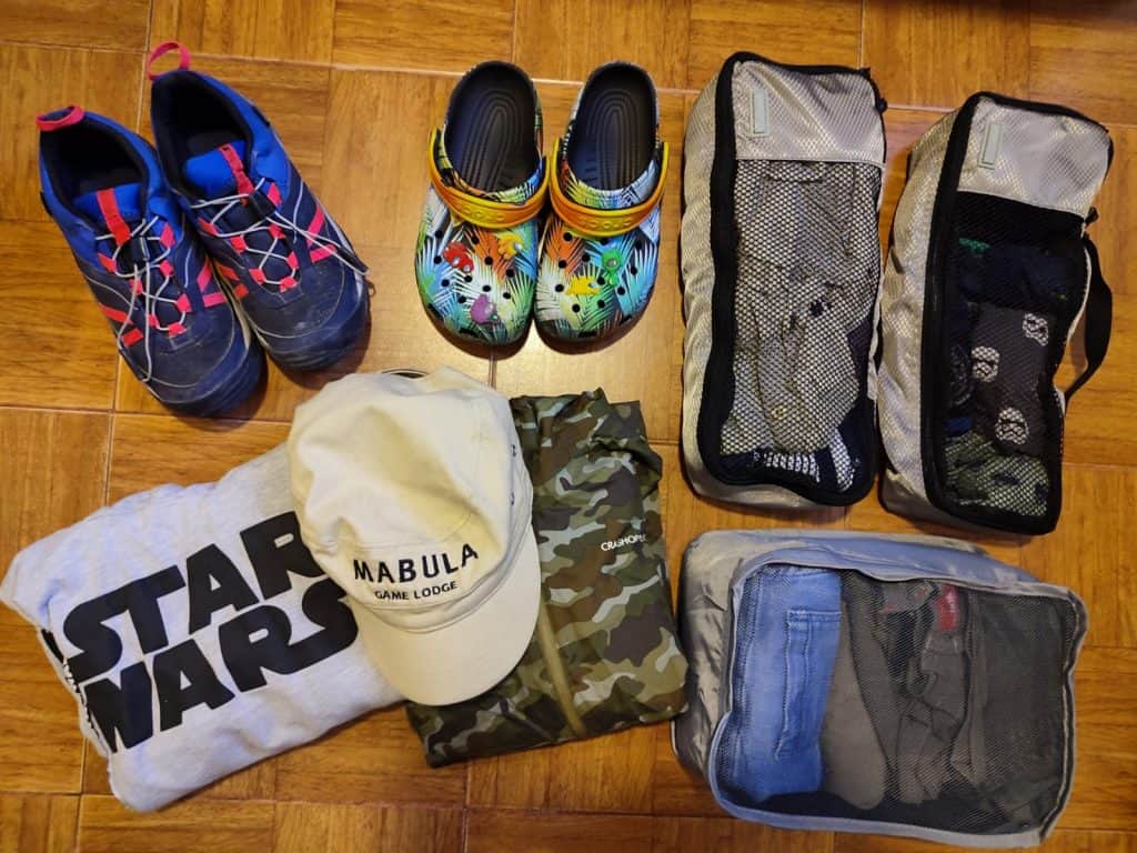 Kids clothes lie on wooden floor in packing cubes, alongside a pair of Crocs, a pair of blue hiking trainers and a cream cap