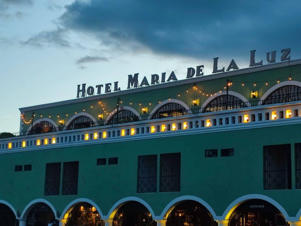 Image of a long colonial style building at sunset, with the lettering 'Hotel Maria de la Luz' across the top of it. Fairy lights are strung along the upper level and the building has archways along it.