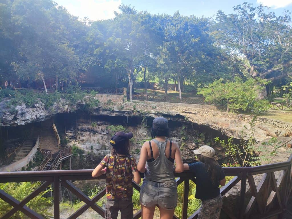 Woman and two children stand at the top of an underground cenote looking down past the wooden railings. 