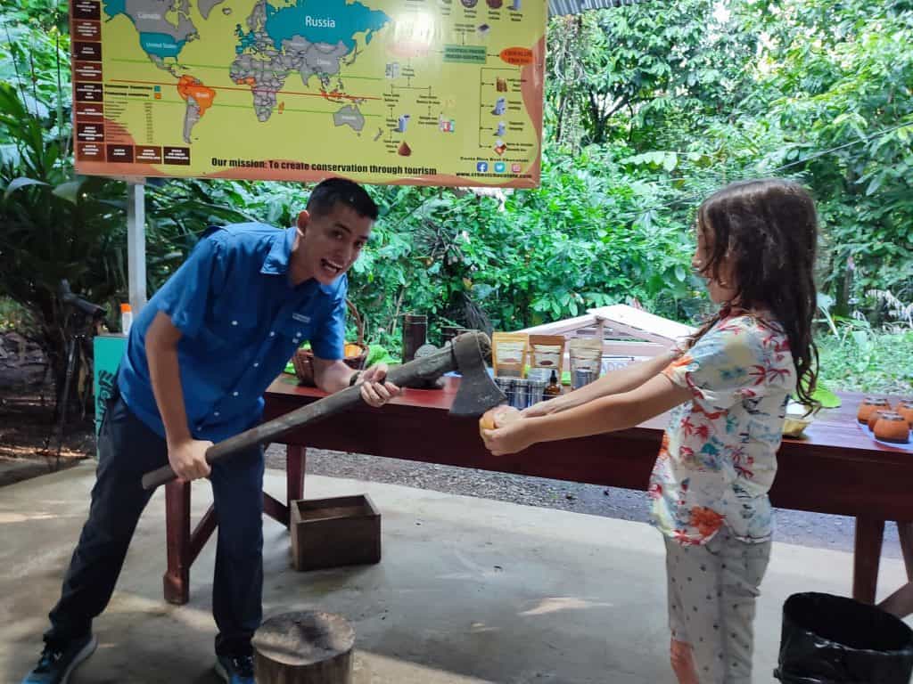 Boy holds cacao pod out in front if him while tour guide holds an axe as if to cut it in half. Behind is a world map showing chocolate producing regions