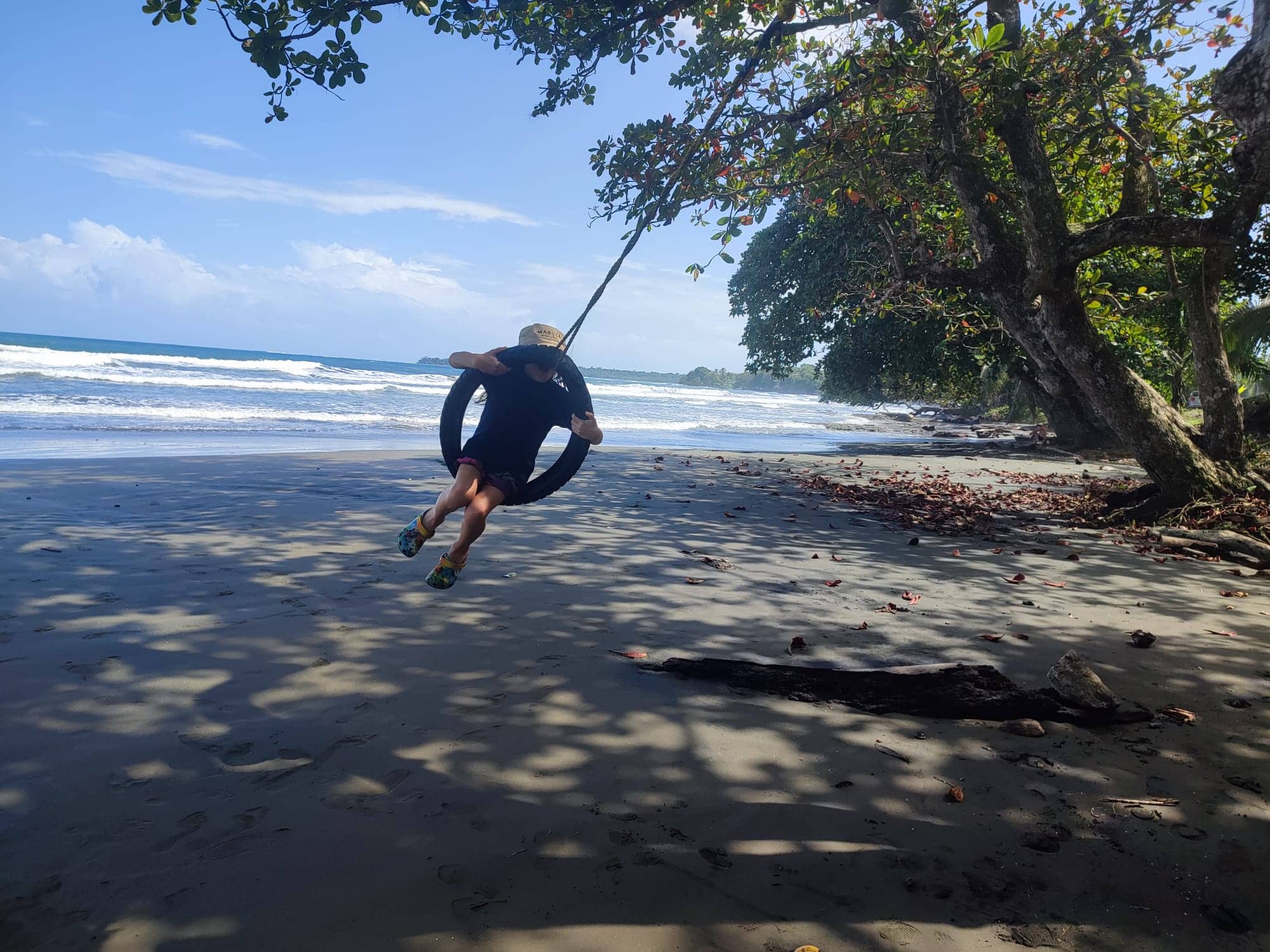 Young boy in swimgear swings on a rubber tyre swing hanging from a rope on the beach. Waves are rolling in in the the background and trees reach over the beach from the right of the frame.