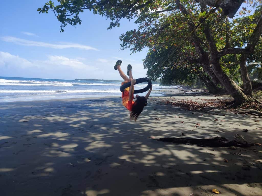 Young boy in swimgear swings high in the air on a rubber tyre swing attached to a rope hanging from a tree on the beach. Waves roll in behind and trees line the edge of the beach