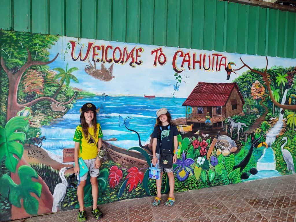 Two young boys stand in front of a brightly coloured mural that states 'Welcome to Cahuita'. The mural shows jungle and a range of animals and bright flowers