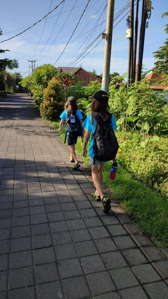 Two youngs boys in school uniform and carrying backpacks walk along a paved road on their way to school. To their left is lush greenery