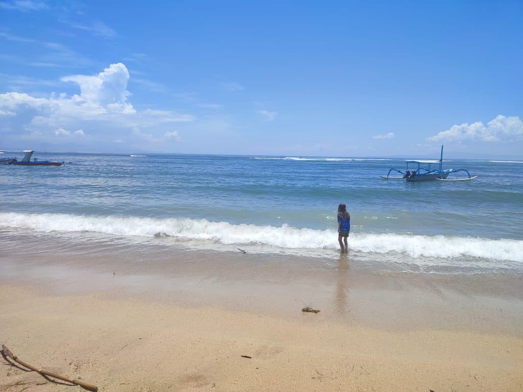 Young boy in swim gear walks through the shallows of the clear blue sea. In the background are two small long boats and waves breaking over an offshore reef