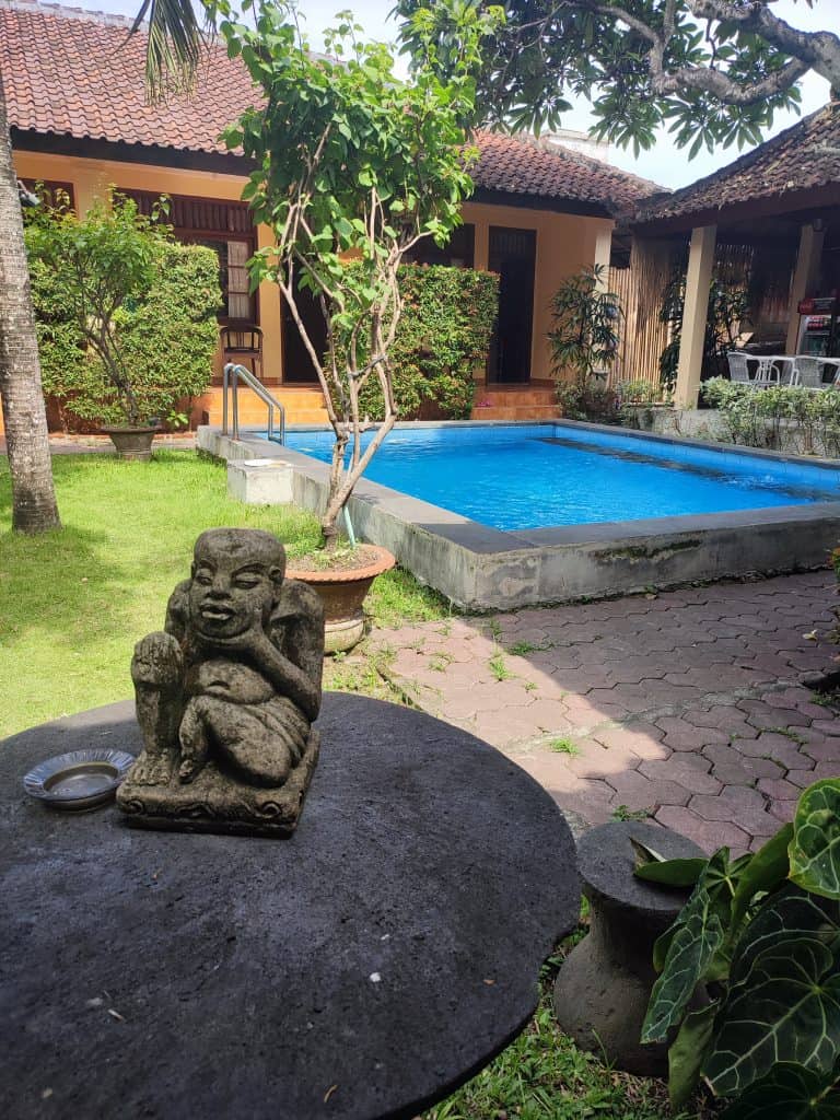 Small guesthouse with rooms surrounding a small swimming pool. A small statue of a sitting person is in the foreground on a table