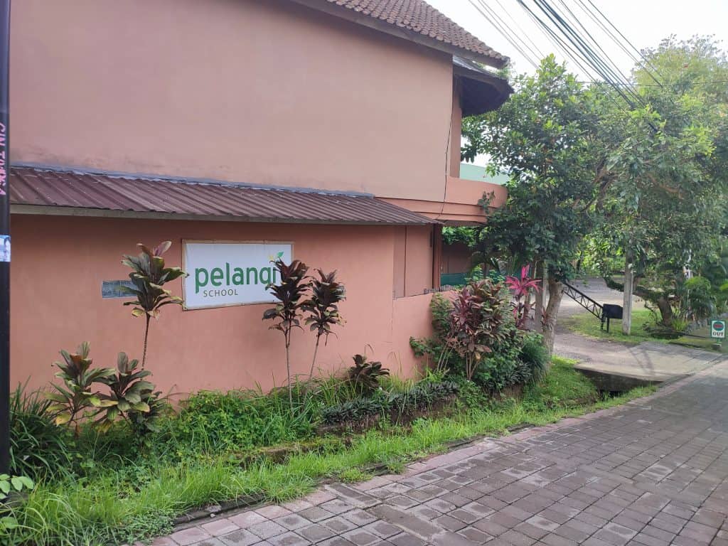 Image of the outside of an international school building where we enrolled our boys as part of our family gap year. The school is painted a coral colour and has a sign stating 'Pelangi School'. In front of the building are several tropical plants