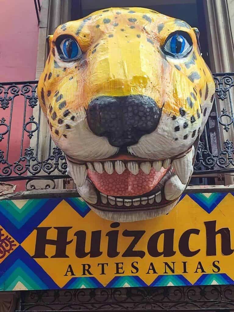 A vibrant sign for Huizache Artesanías in Oaxaca, Mexico, features a colorful jaguar head with striking blue eyes and an open mouth, hanging above a balcony with a decorative iron railing. The sign is adorned with a geometric pattern in blue, green, and yellow