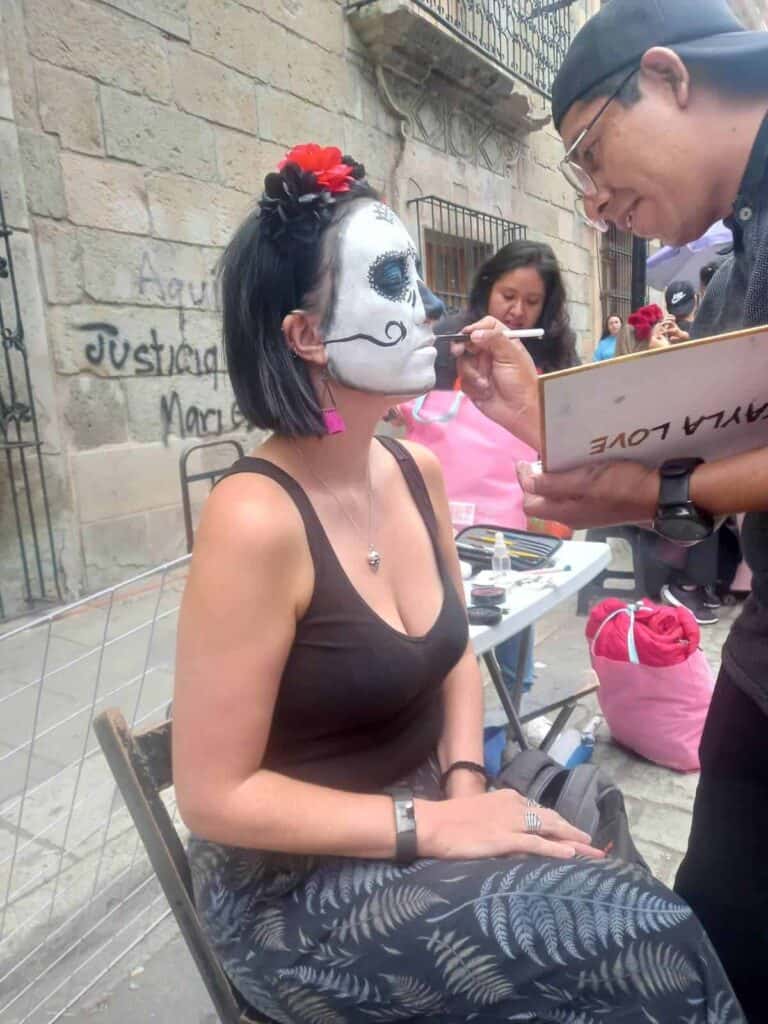 Face painting with a sugar skull design during a Day of the Dead celebration in Oaxaca, Mexico. They sit on a chair in an outdoor setting with a stone wall in the background. A table nearby holds art supplies, and the Huizache Artesanías sign with a jaguar head is visible above.
