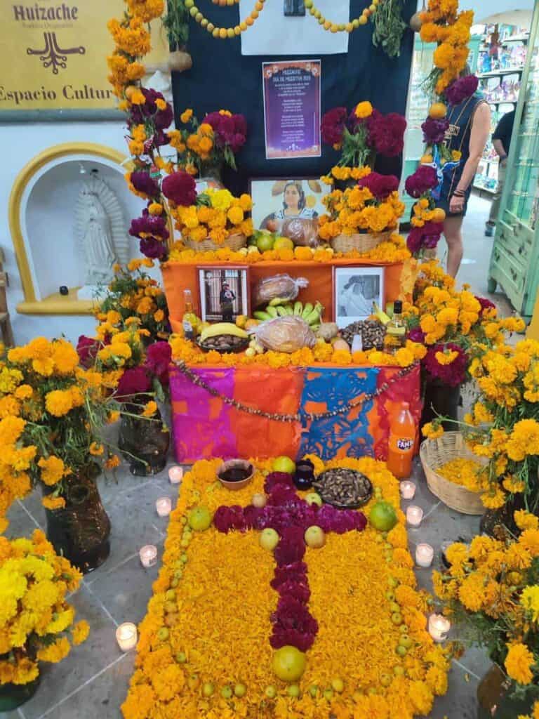 An ornate Day of the Dead decoration in Oaxaca, Mexico, features a doorway framed with marigold flowers, pink celosia blooms, and stacks of gray skulls. A large straw sunburst with a skull at its centre hangs above, creating a festive and vibrant entrance.