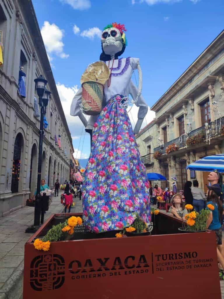 A towering skeleton figure in a floral dress and hat stands in a public square in Oaxaca, Mexico, as part of a Day of the Dead celebration. The display, marked with a sign from Oaxaca Turismo, is surrounded by marigold flowers and set against a backdrop of historic buildings and a lively crowd.