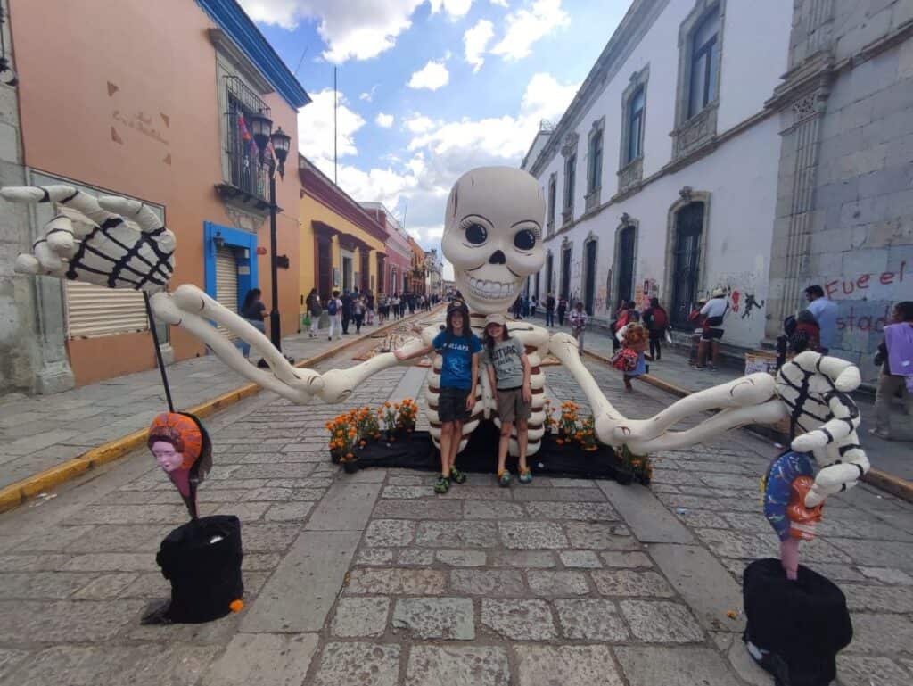 Two children pose with a large skeleton during a Day of the Dead event in Oaxaca, Mexico. The skeleton is surrounded by marigold flowers, and the street is lined with historic buildings and other festive decorations, capturing the lively atmosphere of the celebration