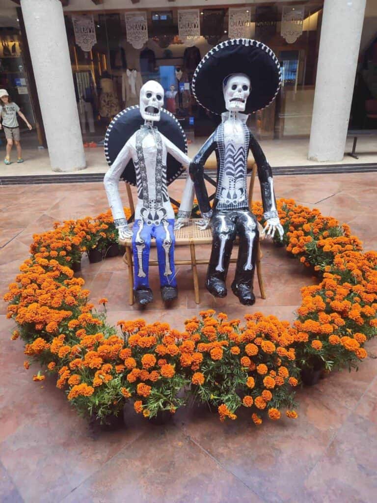 Two skeleton figures dressed in traditional Mexican clothing sit on a bench during a Day of the Dead celebration in Oaxaca, Mexico. They are surrounded by a ring of marigold flowers, with papel picado banners hanging in the background, adding to the festive atmosphere.