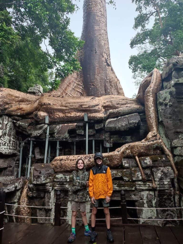 Two boys in raincoats stand in from of a giant tree root at Ta Prohm temple, Angkor Wat on a family visit