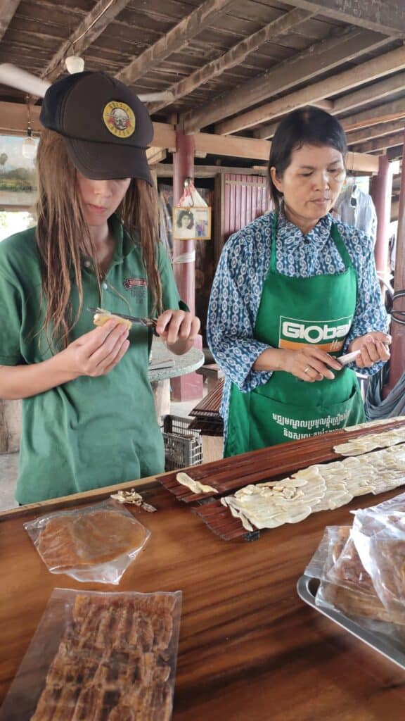 Boy slices banana with Cambodian woman. Wooden board is on the table ready for them to be laid on to dry in the sun in Battambang