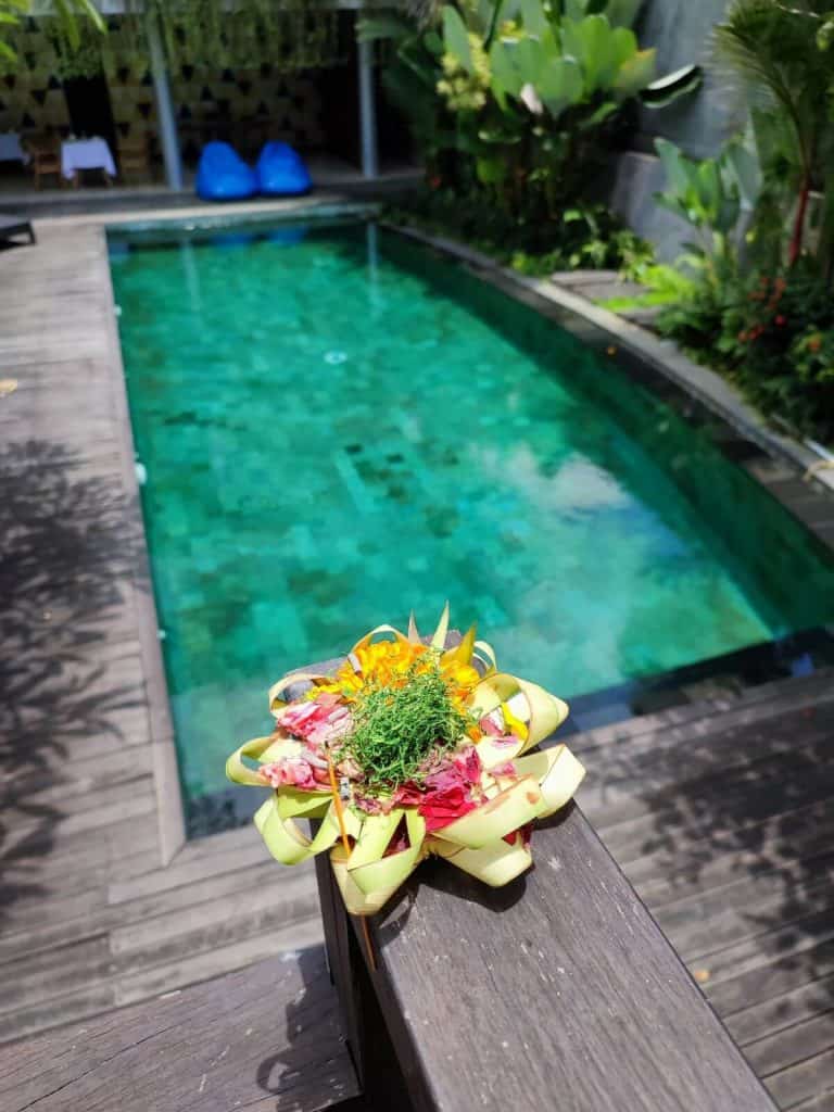 A vibrant Balinese Canang Sari offering with colorful flowers and woven palm leaves rests on a wooden railing overlooking the emerald-green pool at Hotel Athaya in Ubud. The lush tropical surroundings and serene water create a peaceful retreat.