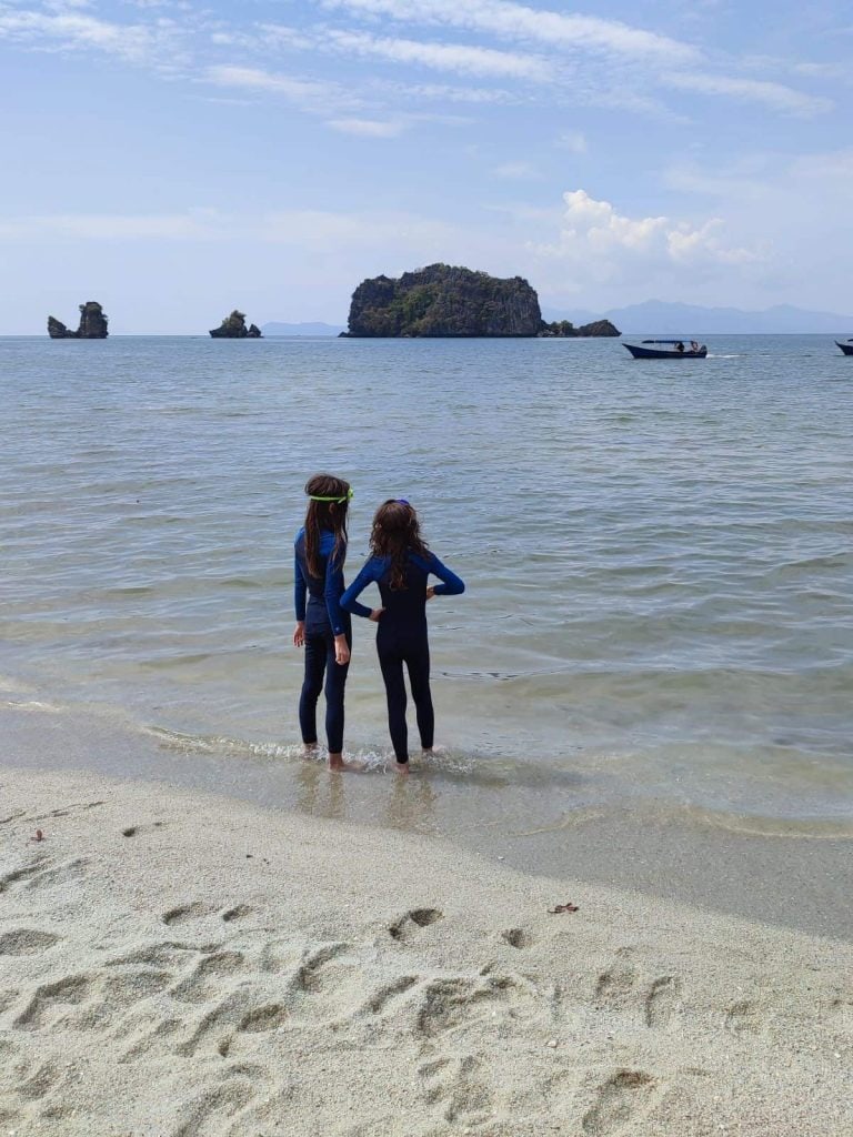 Two boys in full length blue swimsuits stand at the water's edge on a beach overlooking some rock formations out to see.