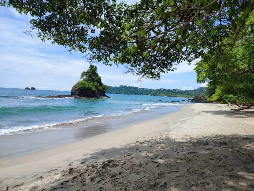 Stretch of white sand, aqua sea with gentle waves and jungle background. Located just offshore is a rock formation covered in green foliage. At Manuel Antonio beach inside the National Park in Costa Rica.