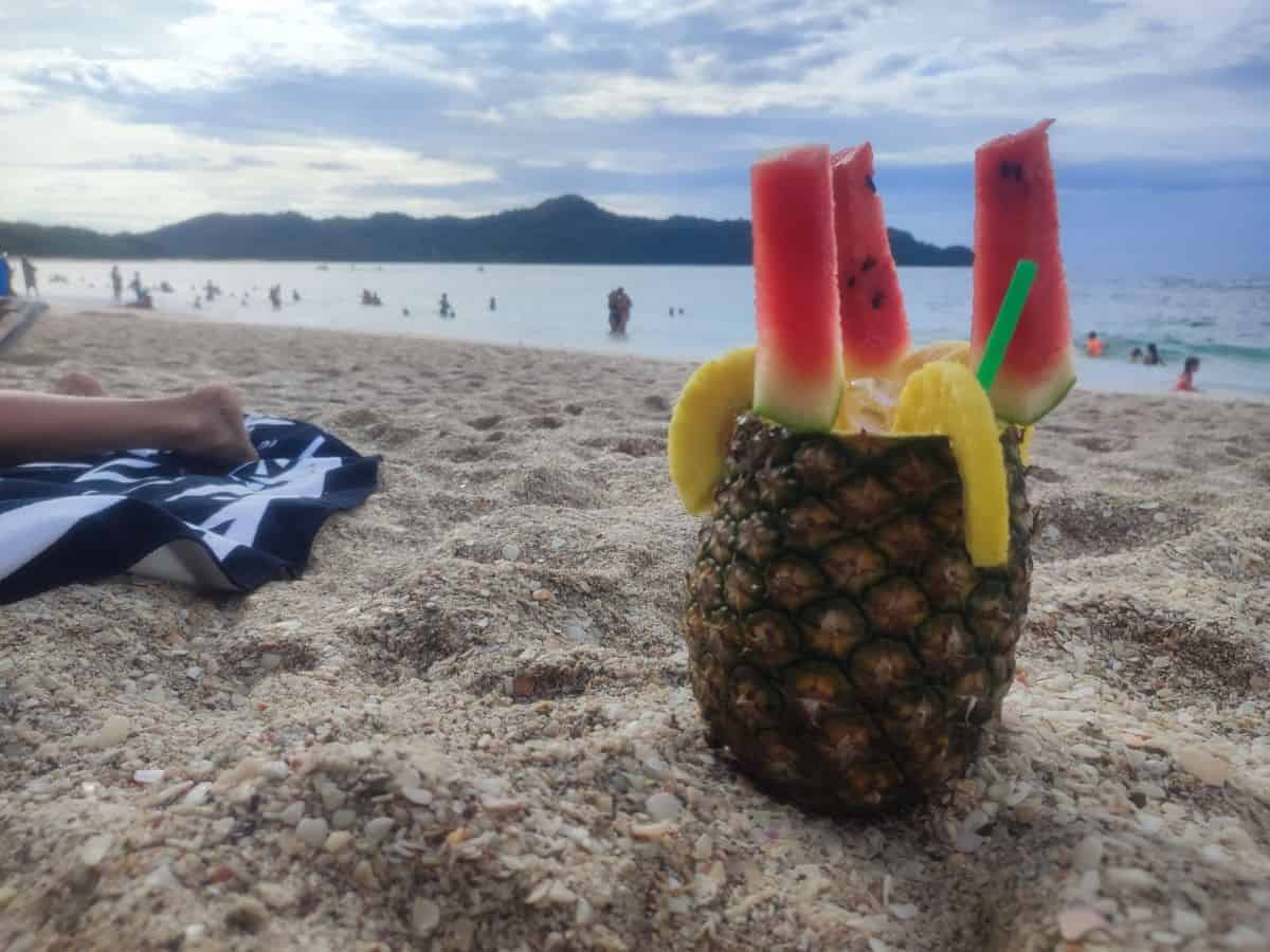 A cocktail in a pineapple with watermelon slices in the top on Playa Conchal beach in Costa Rica. In the background is the blue sea with a number of people wading and swimming in it.