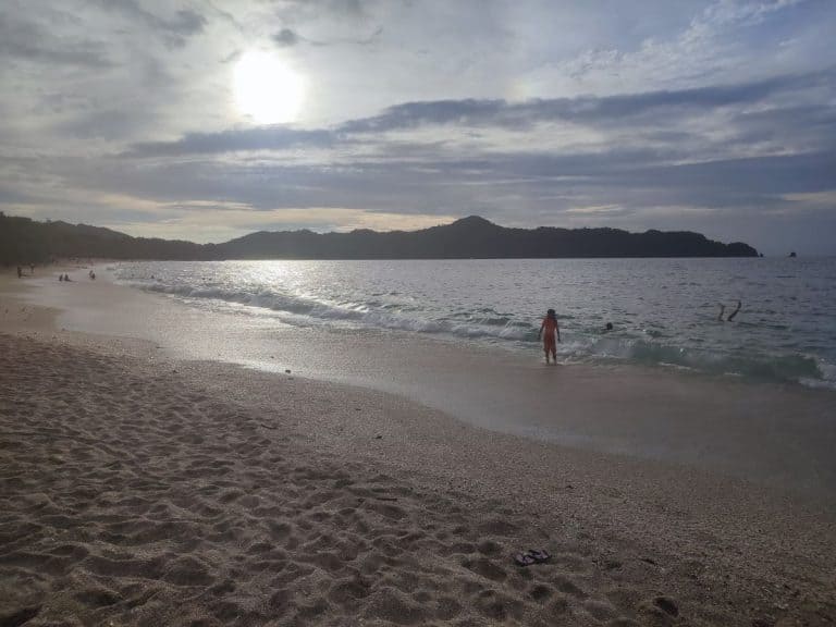 A family enjoys a sunny day at a Costa Rican beach, with kids playing in the shallow waves and parents relaxing on the sand, ideal for Costa Rica travel with kids.