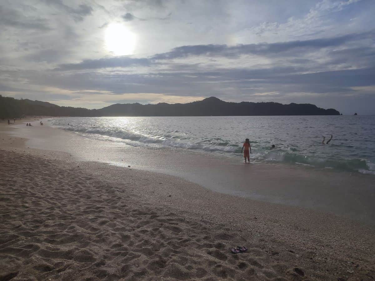 A family enjoys a sunny day at a Costa Rican beach, with kids playing in the shallow waves and parents relaxing on the sand, ideal for Costa Rica travel with kids.