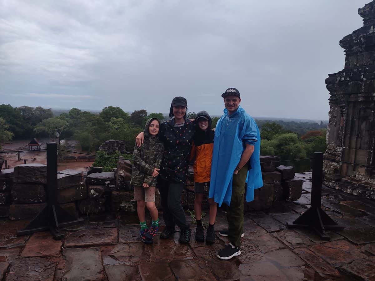 Family of four in raincoats stand at the top of a temple at Angkor Wat with the Cambodian countryside in the background.