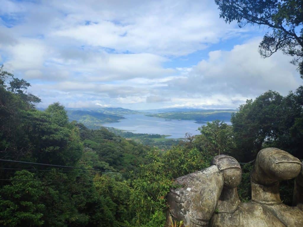 Giant hand sculpture in the foreground with views out over Arenal Lake and the lush green jungle beyond in Arenal/La Fortuna, Costa Rica.