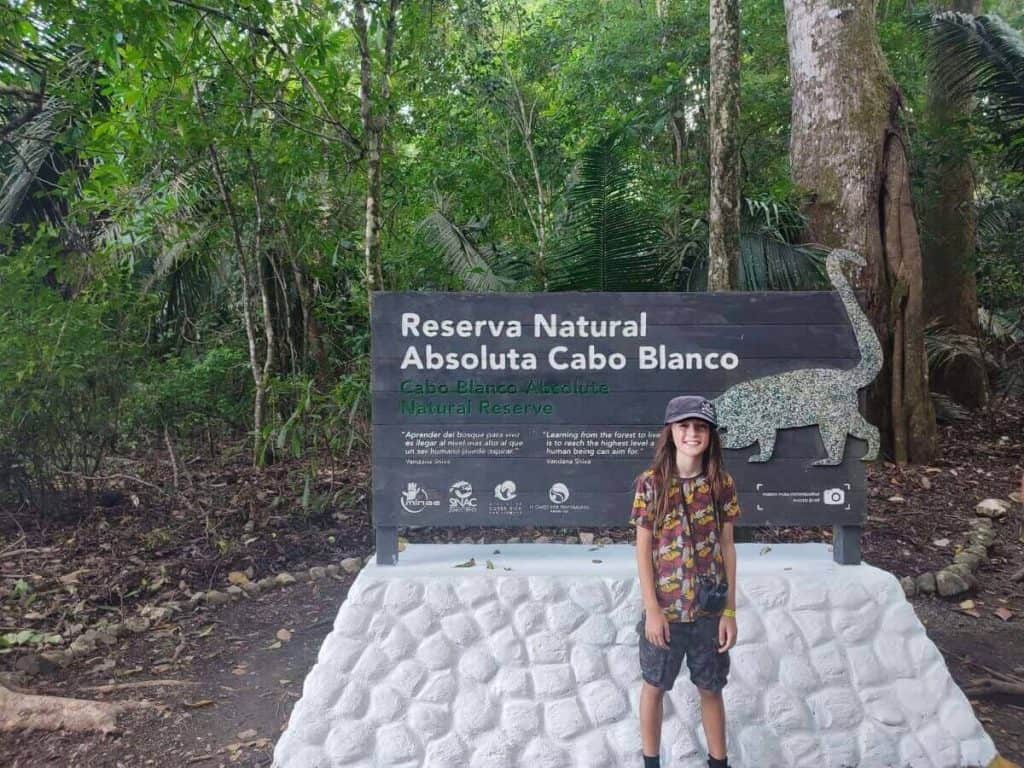 A child stands in front of the Reserva Natural Absoluta Cabo Blanco sign in Costa Rica, ready for a nature adventure, highlighting Costa Rica travel with kids.