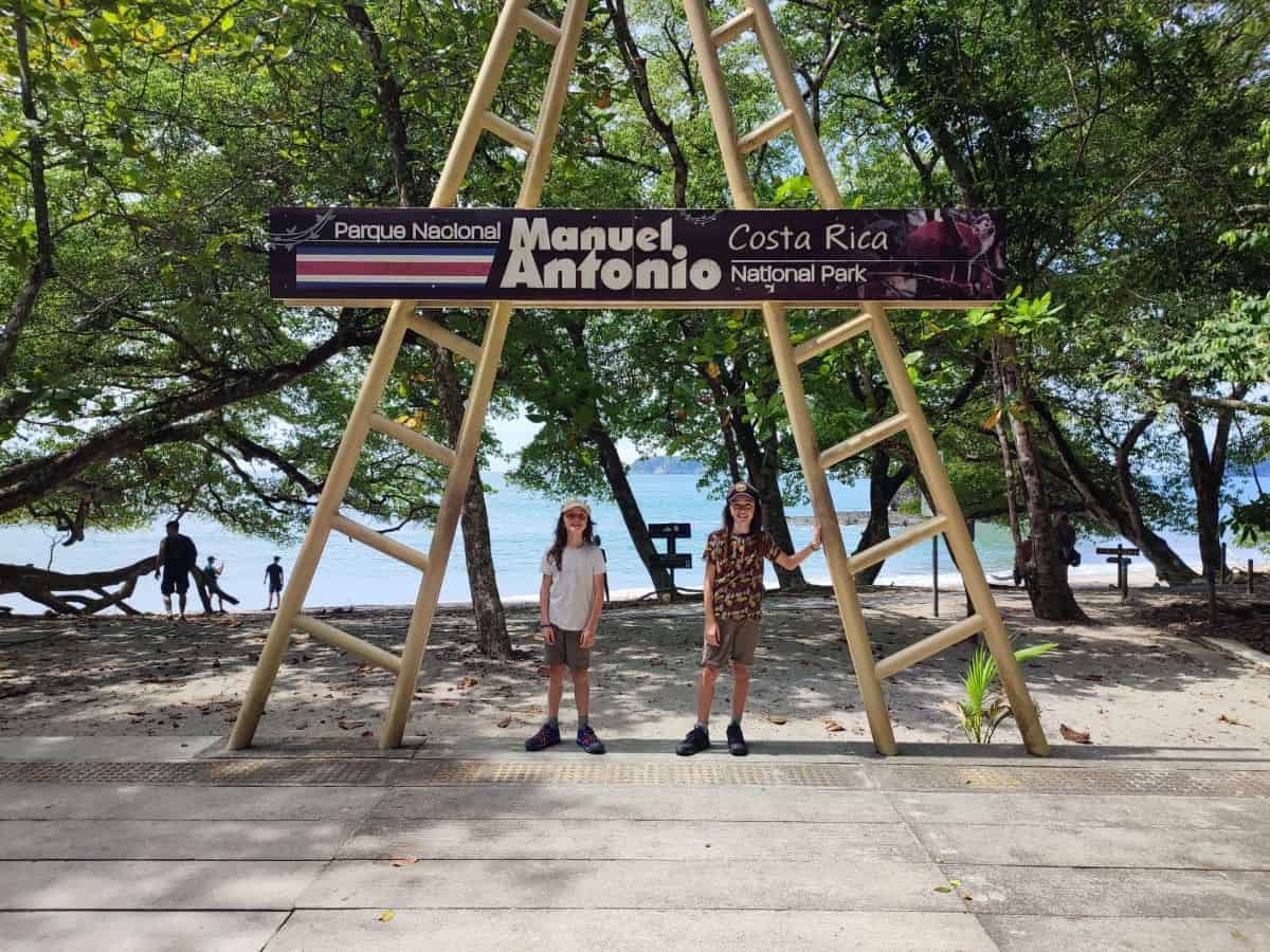 Two boys stand beneath a wooden sign that says 'Parque Nacional Manuel Antonio' in Costa Rica. behind is a white sand beach and the sea.