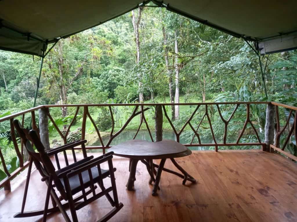 A wooden rocking chair and small carved table sit on a wooden deck under canvas, looking out over a natural pond at Rafiki Safari Lodge Costa Rica.