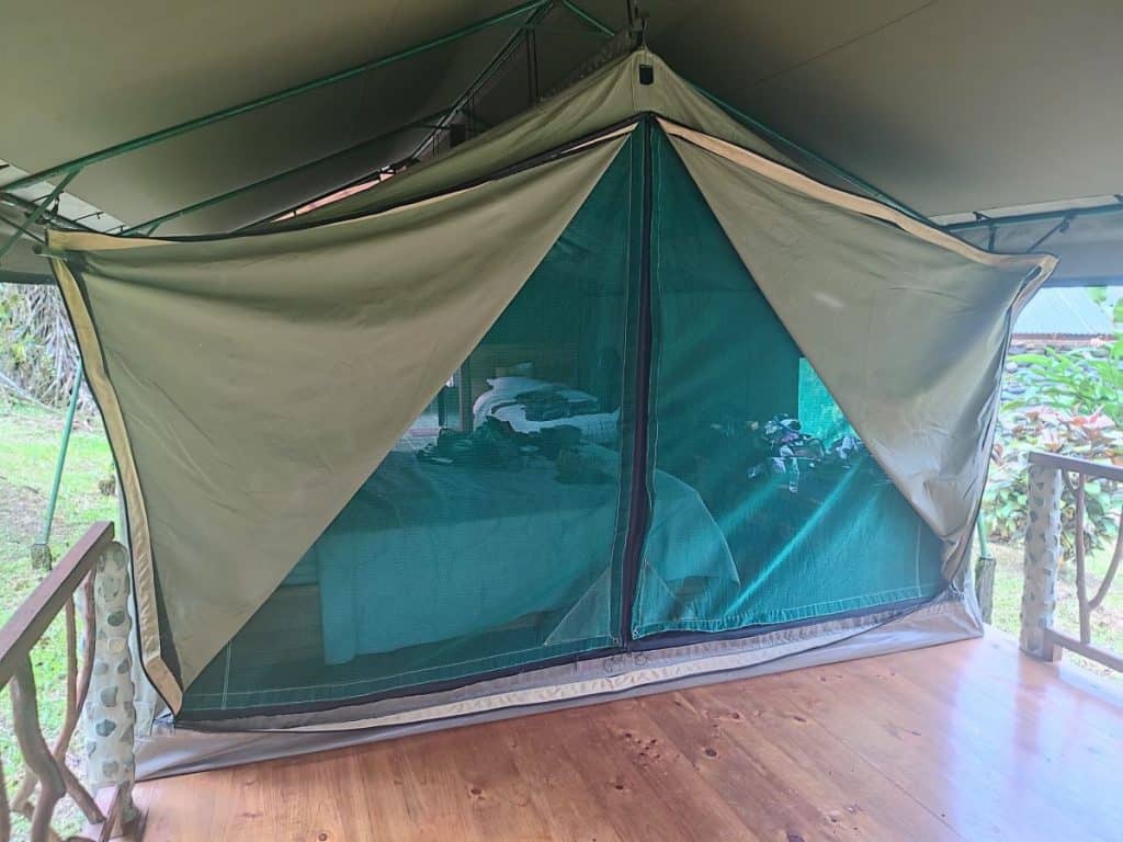 The outside of a large safari tent at Rafiki Safari Lodge, with the front flaps opens so you can see the bed inside. To the front is a wooden decked area.