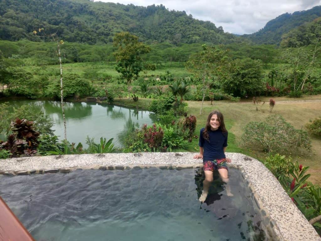 View across a small pool with a young boy sitting on the edge of it, out across the plains of Costa Rica and the jungle beyond at Rafiki Safari Lodge, Costa Rica.