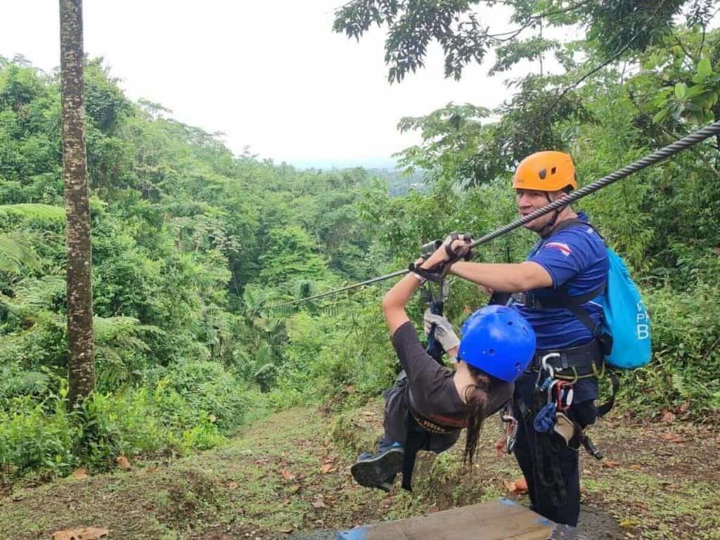 Child attached to a zipline leans back ready for take off, supported by a guide. The zipline descends into the Costa Rica rainforest in La Fortuna, Costa Rica.