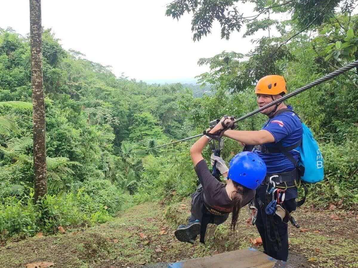 Child attached to a zipline leans back ready for take off, supported by a guide. The zipline descends into the Costa Rica rainforest in La Fortuna, Costa Rica.