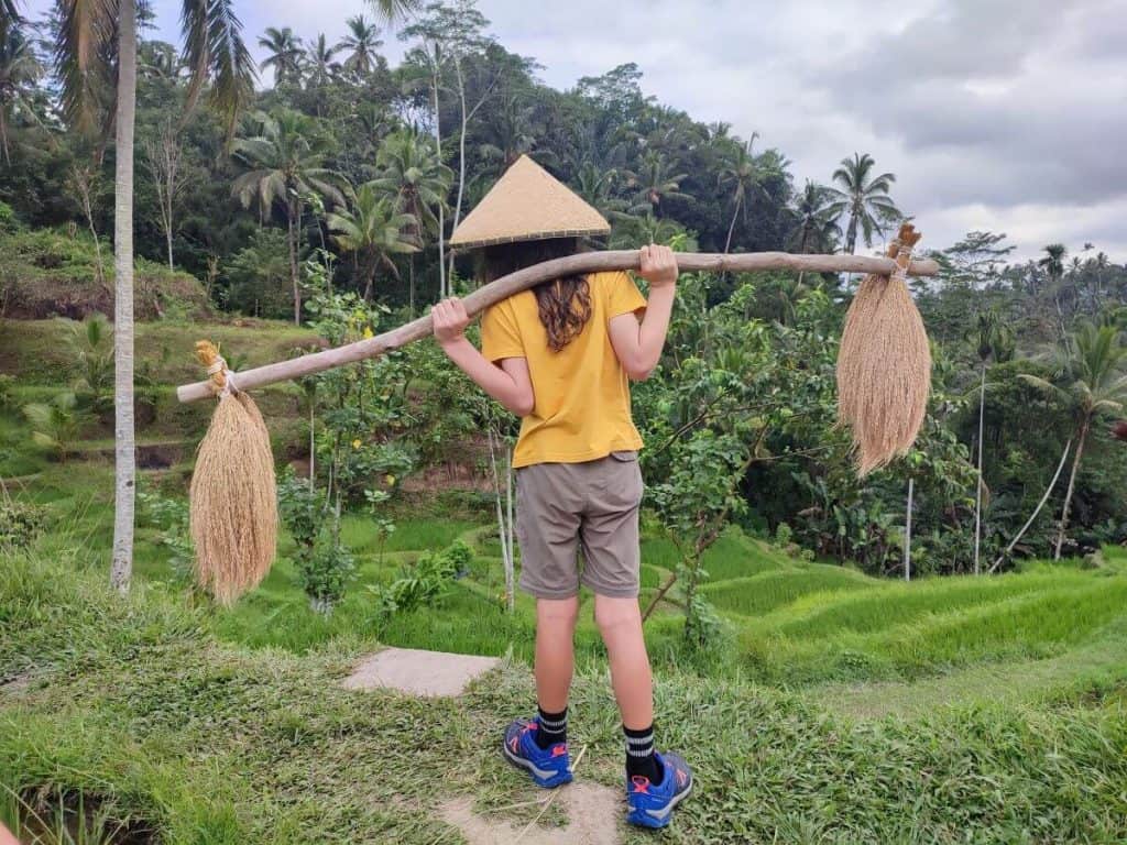 A child wearing a conical hat and holding a traditional carrying pole in the terraced rice fields of Ubud, Bali
