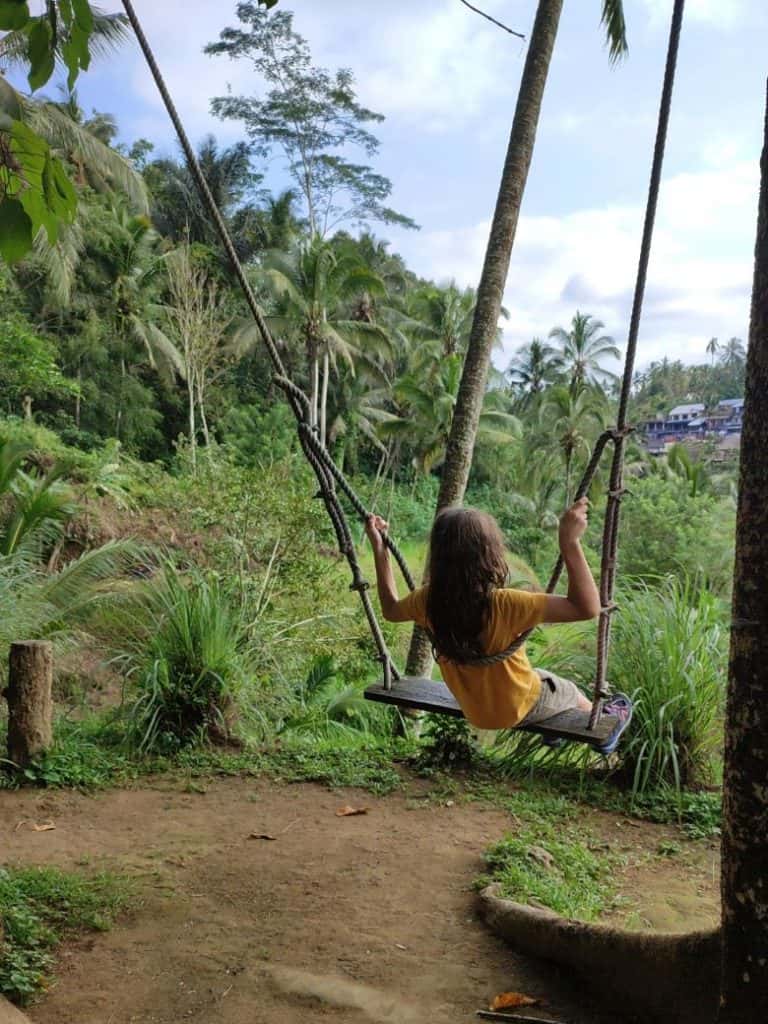A child on a wooden swing overlooking the stunning terraced rice fields in Ubud, Bali, a must-see for families visiting the best rice fields in Ubud