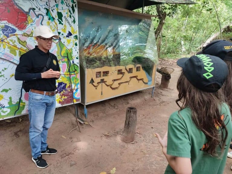 Tour guide stands in front of a model of the Cu Chi Tunnel complex in Vietnam while two boys listen on.