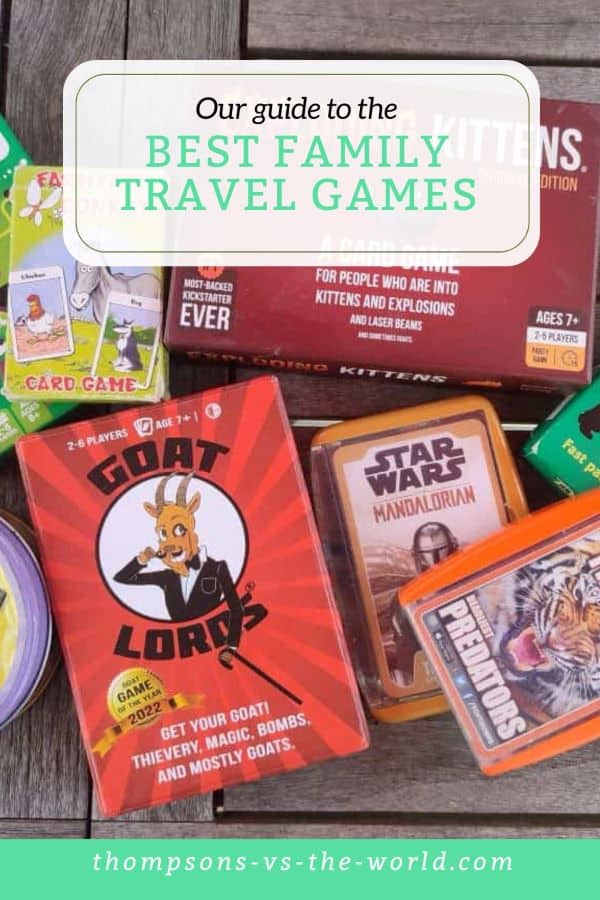 Various family travel games sit in a pile on a wooden table, including Goat Lords, Top Trumps and Exploding Kittens. Text overlay reads 'Our Guide to the Best Family Travel Games' Save this pin for later!