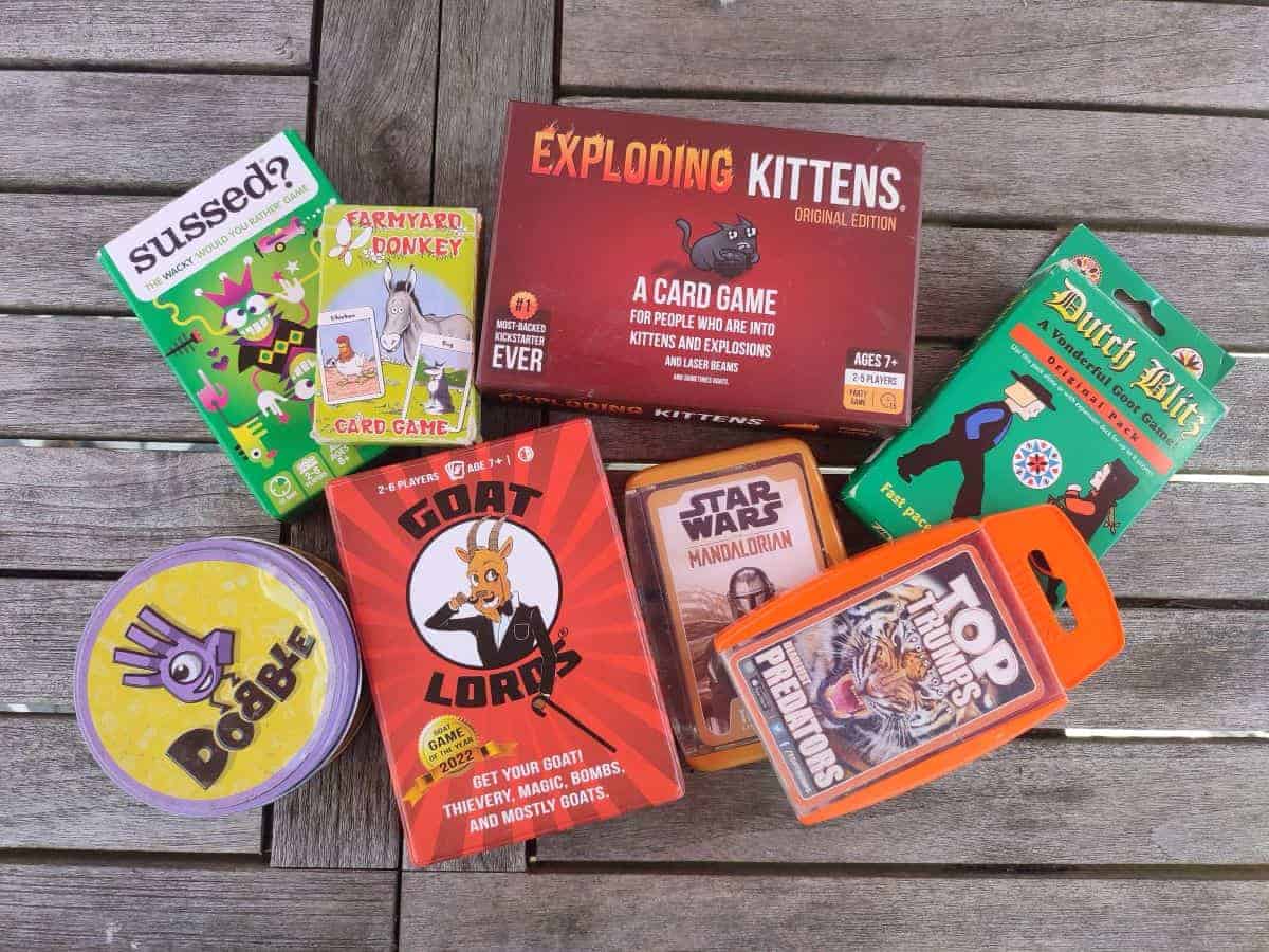 A pile of family travel games sits on a wooden table, including Exploding Kittens, Goat Loads, Dobble, Top Trumps, Dutch Blitz and Sussed. Some of our top recommendations for the best family travel games!