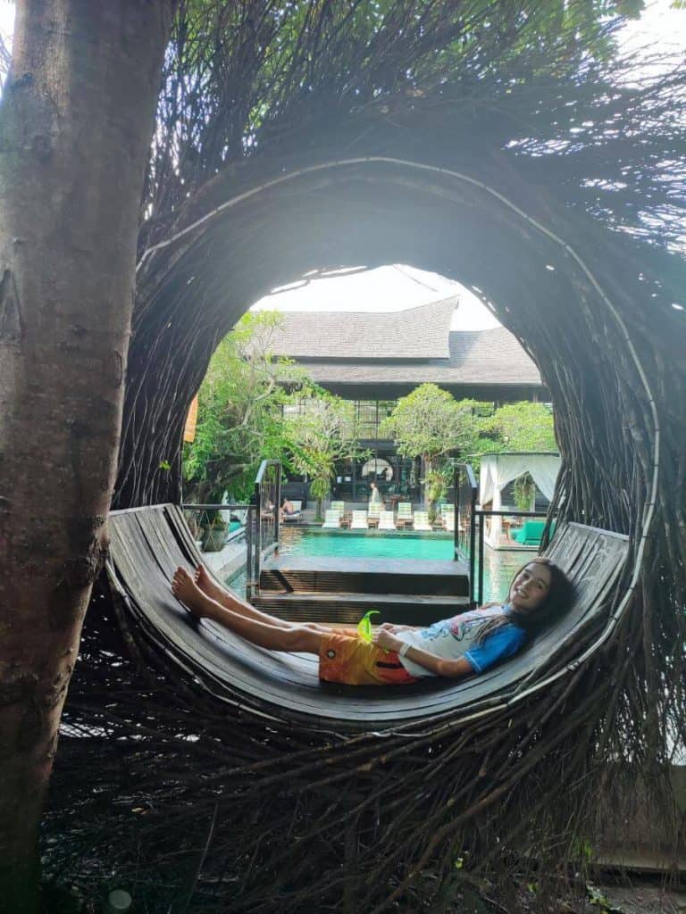 A child relaxes in a woven hammock framed by a circular tree structure at Titi Batu Ubud, with a pool and resort in the back.