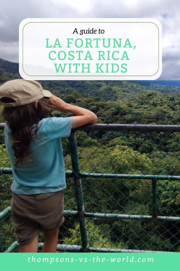 Scenic view of a lush green jungle in La Fortuna, Costa Rica, with a child in a cap and blue shirt leaning on a railing, overlooking the verdant landscape under a cloudy sky. Text overlay reads "A guide to LA FORTUNA, COSTA RICA WITH KIDS" in green and white. Perfect for families planning a trip to La Fortuna with kids! Save this pin to your Pinterest board for later reference.