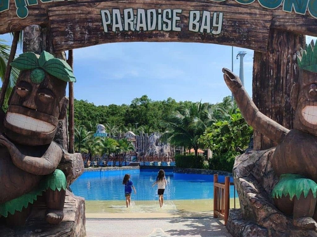 Two children walk toward a bright blue pool framed by a large rustic wooden archway that reads Paradise Bay, with smiling tiki-style statues on each side. Lush greenery and tropical scenery surround the water park, capturing a playful family atmosphere. This image highlights one of the fun things to do in Phu Quoc Vietnam - Aquatopia Waterpark.