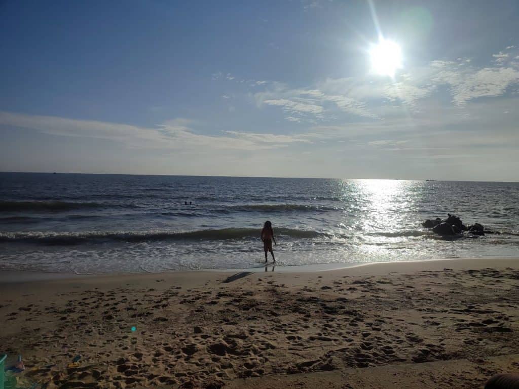 Young boy stands in the shallows of the ocean as the evening sun starts to go down. There is a long expanse of yellow sand in the foreground and the light from the sun across the small waves in the sea.